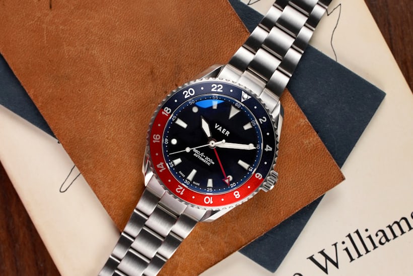 A GMT watch with a red and blue bezel laying down on a piece of brown leather.