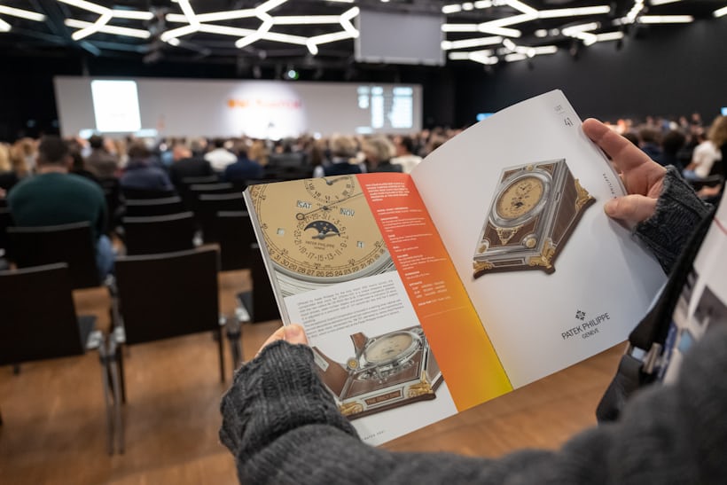 A man holds the Only Watch 2021 catalog up and is reading the page dedicated to the Patek Philippe contribution while the Only Watch auction takes place in the background.