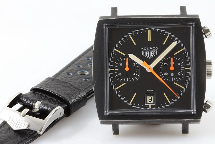 The dial of a black Heuer Monaco with a black strap in the background