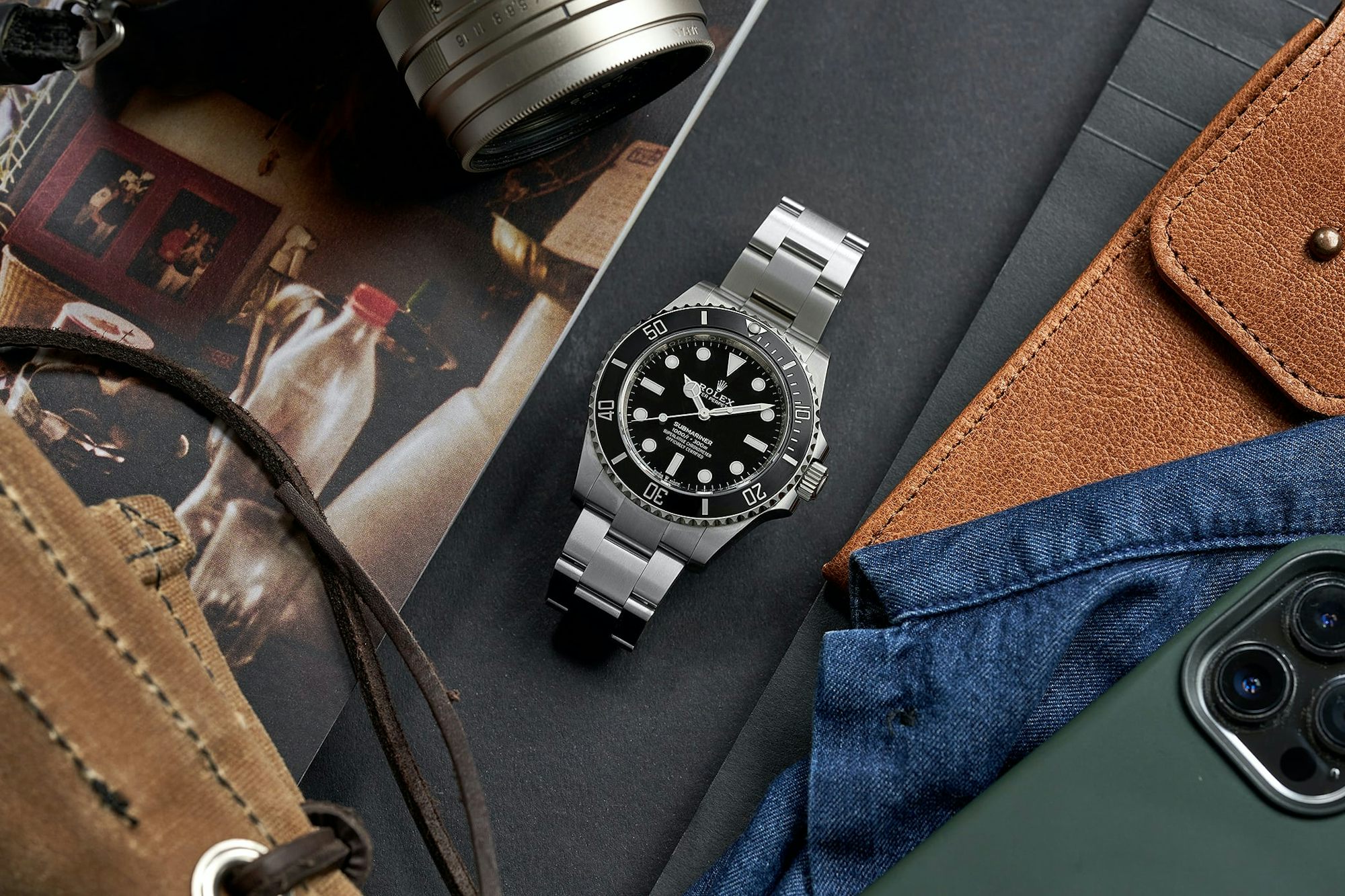 Rolex Submariner next to iPhone, camera, and other desktop items.