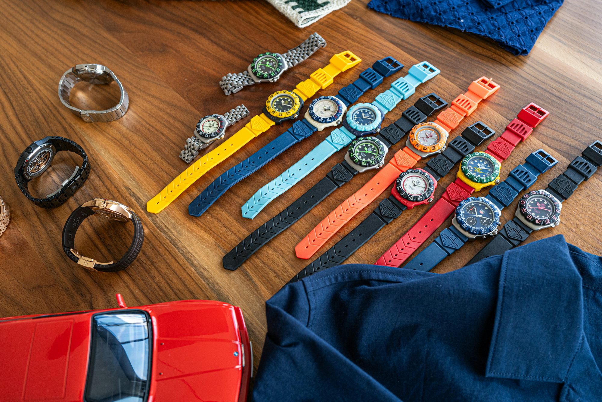 A collection of colorful watches in different styles and metals laid out on a wooden table next to some fabric and a model car
