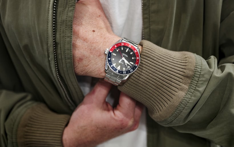 A wrist shot of a man wearing a GMT watch with a red and blue bezel. He is reaching into the inner pocket of his green jacket.