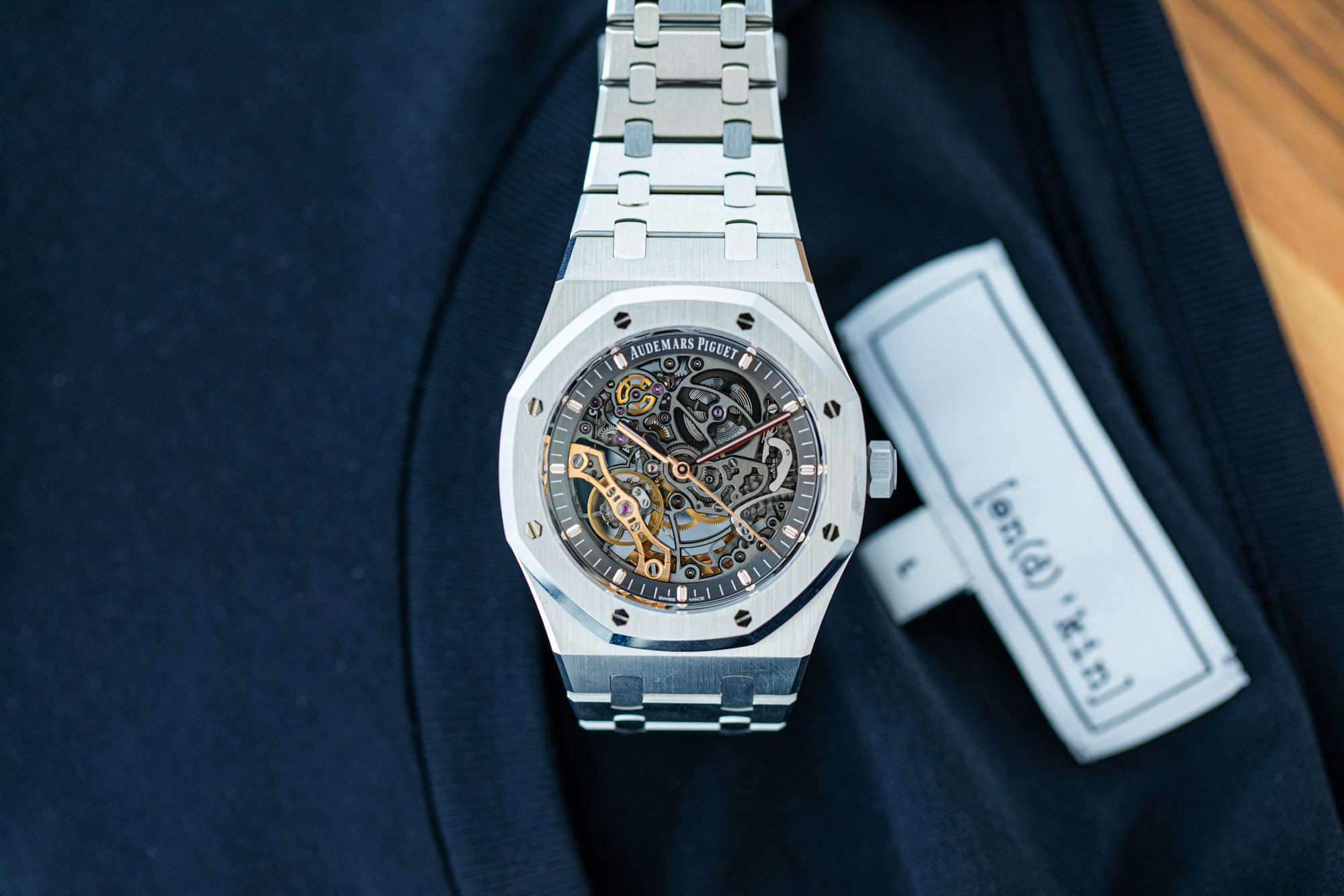 Audemars Piguet Double Balance Wheel in Steel on a background with text on a shirt tag that reads "[en(d) 'kin]" size L