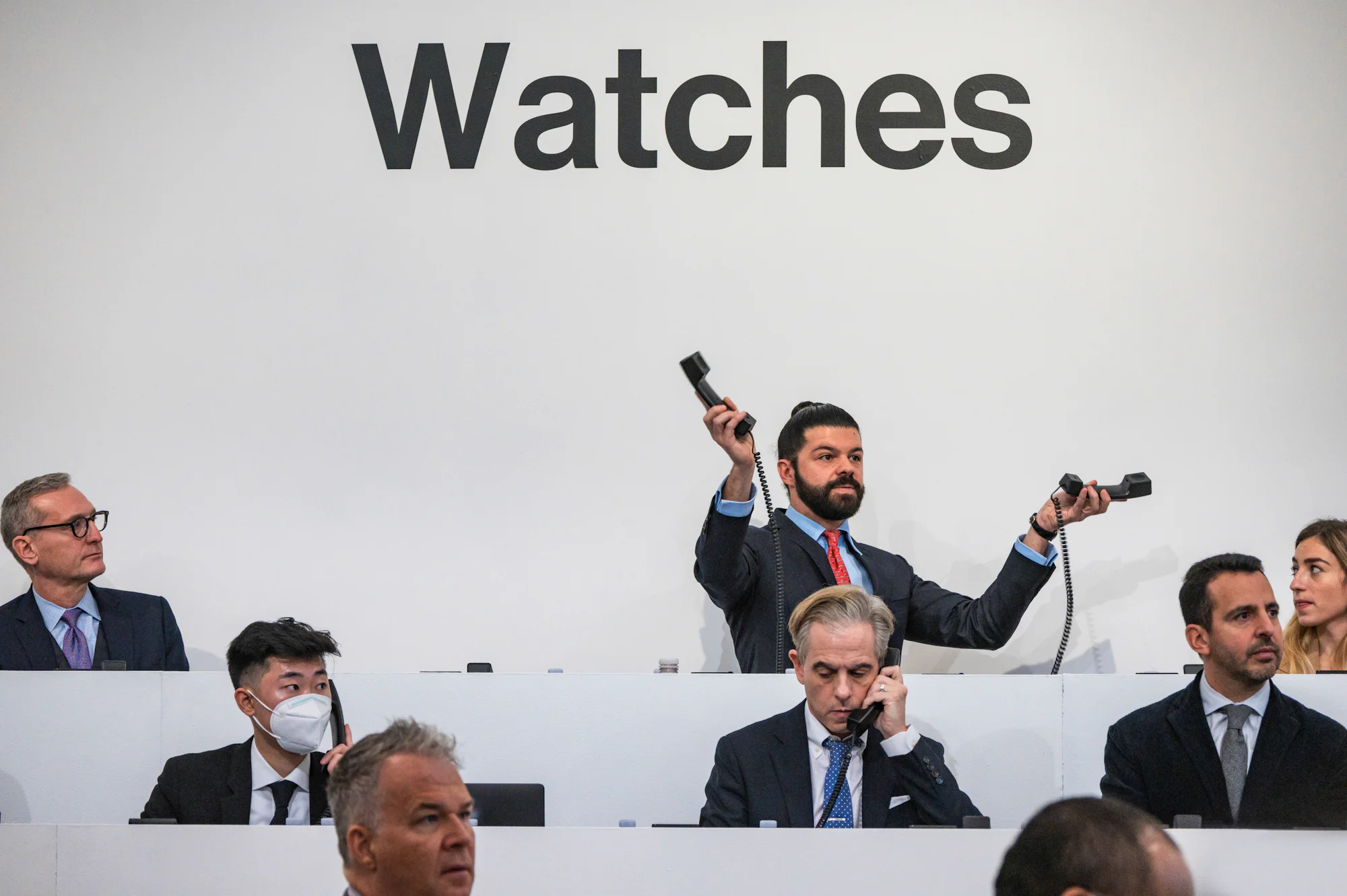 A man with a beard and man bun wearing a suit stands with one phone in each hand.