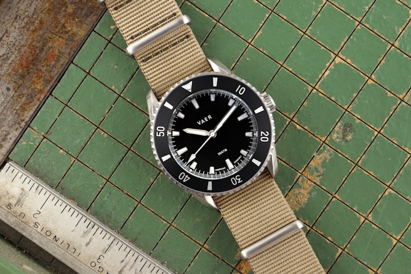 A black dial dive watch on a tan NATO-style strap lays down on a faded green background.
