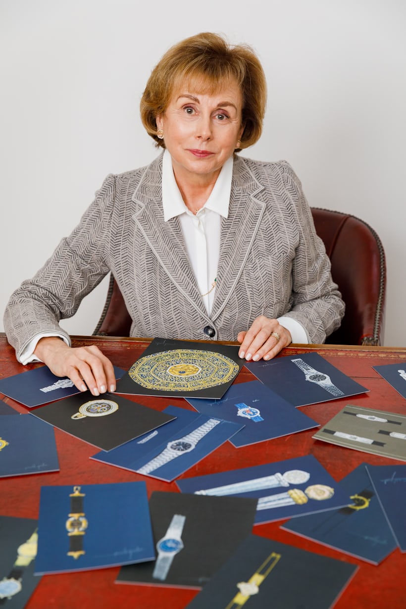 A woman looks at the camera and sits in front of a red table with various sketches of Genta watches.