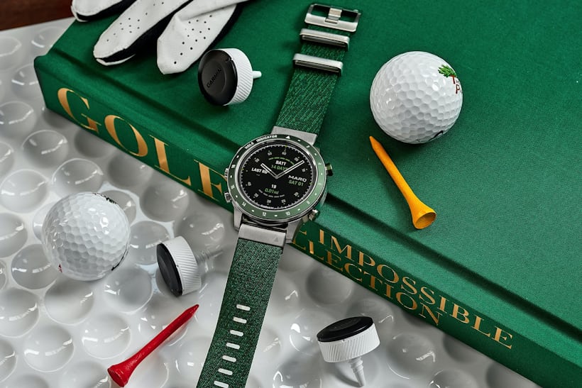 The Garmin MARQ Golfer 46mm laying atop Assouline's "Golf: The Impossible Collection" with assorted golf paraphernalia laying around it.